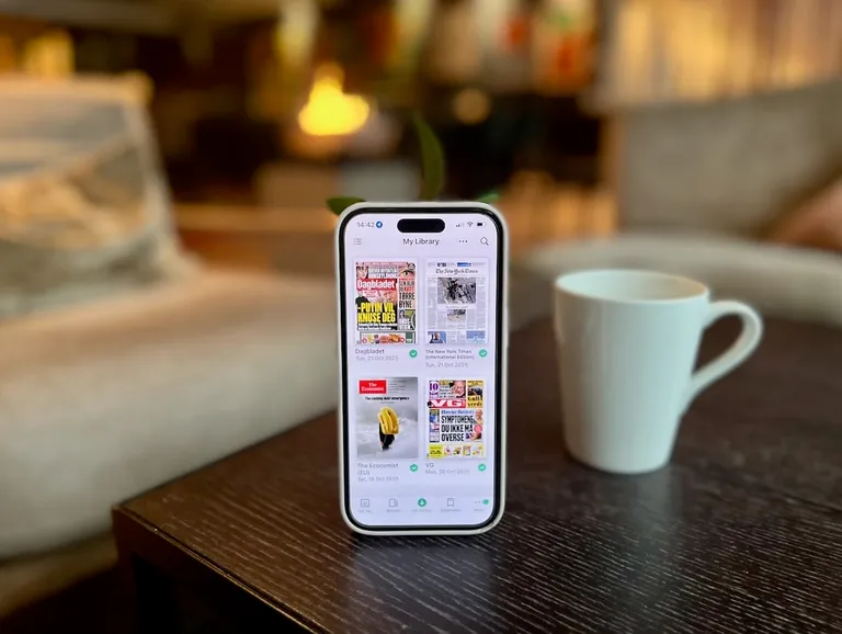 Bilde av en mobiltelefon med PressReader-appen åpen, i bakgrunnen vises en kaffekopp og en peis